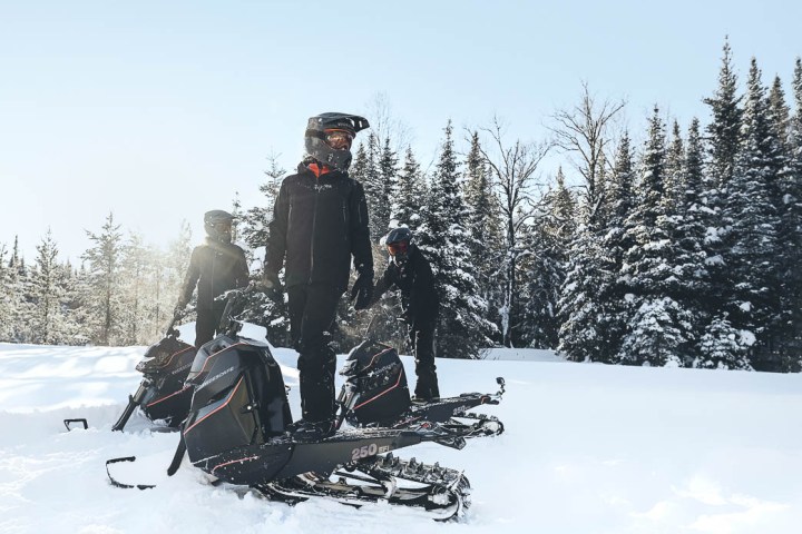 Three snowmobilers in helmets standing in snowy forest landscape with snowmobiles.